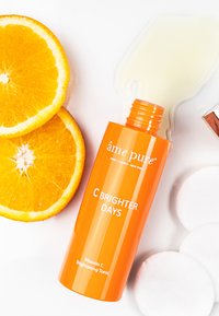 Lysende orange flaske mærket "C BRIGHTER DAYS" med hvid tekst, indeholdende en gul væske, omgivet af appelsinskiver og vatrondeller.
