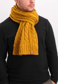 Yellow knitted scarf with a cable pattern, thick ribbed edge, and a leather label. Worn over a textured black sweater.