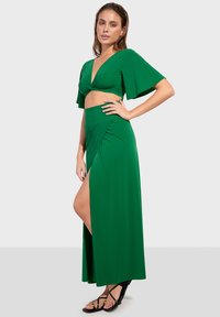 Set verde in due pezzi composto da una maglietta corta con scollatura a V e maniche a strascico, accompagnata da una lunga gonna drappeggiata con uno spacco alto fino alla coscia.