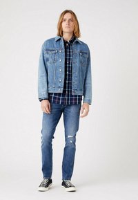 Jaqueta de ganga azul clara com dois bolsos no peito, fecho de botão, usada sobre uma camisa xadrez escura. Combinada com jeans azul desbotado e sapatilhas pretas.