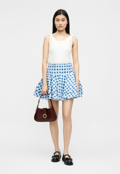 Νεαρή γυναίκα που φοράει λευκό αμάνικο τοπ, μπλε και λευκή καρό φούστα με βολάν, μαύρα παπούτσια, κρατώντας σκούρο καφέ τσάντα, στέκεται μπροστά σε λευκό φόντο.