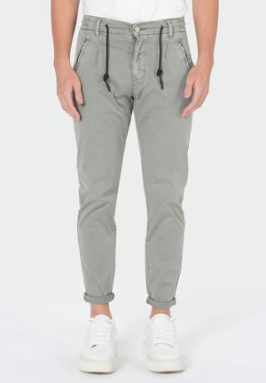 Mann trägt hellgraue Freizeithose mit umgeschlagenen Säumen, schwarzen Kordeln und weißen Sneakers, steht vor einem schlichten Hintergrund.