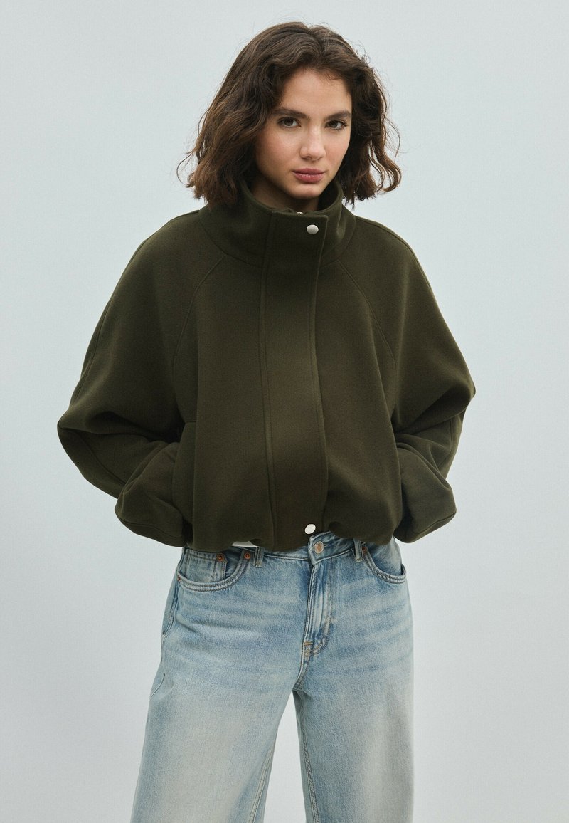 Jeune femme aux cheveux courts et ondulés porte une veste à col montant vert olive foncé et un jean taille haute bleu clair, les mains dans les poches.