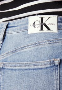 Jeans de mezclilla azul claro con una etiqueta blanca que lleva el logo "Calvin Klein Jeans", costuras en el bolsillo trasero y un diseño clásico de cinco bolsillos.