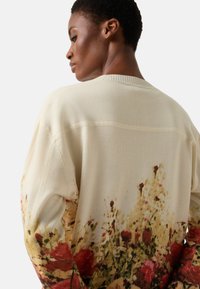 Kremowy sweter z kwiatowym nadrukiem u dołu w odcieniach czerwieni i żółci, miękka tekstura oraz ściągacz przy dekolcie.