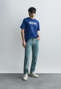 Μπλε γραφικό μπλουζάκι με το λογότυπο "MOSCHINO", ανοιχτόχρωμο τζιν και λευκά αθλητικά παπούτσια· το μοντέλο στέκεται μπροστά σε απλό φόντο, φορώντας σκούρα γυαλιά ηλίου.