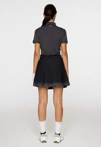 Vrouw die met haar rug naar de camera staat, gekleed in een donkere poloshirt, geplooide rok, witte handschoen, witte sokken en sneakers tegen een effen achtergrond.