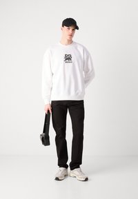 Hvit sweatshirt med svart logo, svarte bukser og grå sneakers. Holder en svart bag. Enkel design med løs passform og rund hals.
