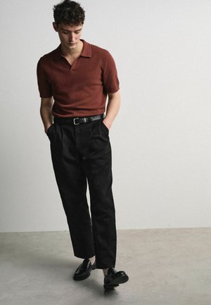 Vyras su garbanotais plaukais, vilkintis sraigės spalvos trumpomis rankovėmis polo marškinėlius, juodą diržą, juodas kelnes ir juodus loafers batus, stovintis su rankomis kišenėse.