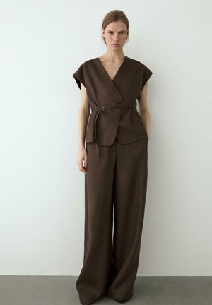 Donna in piedi contro una parete bianca che indossa un top marrone senza maniche con cintura e pantaloni marroni a gamba larga.