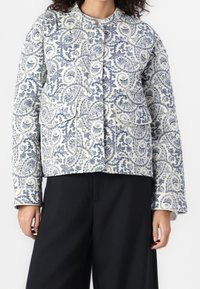 Quiltet jakke i hvid med detaljeret blåt paisley-mønster. Har en rund halsudskæring, frontknaplukning og to påsyede lommer.