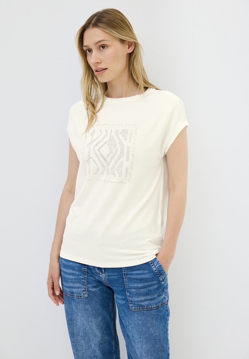 Cremefarvet T-shirt med korte ærmer og grafisk tryk med tekst og abstrakt mønster. Blødt stof, afslappet pasform, kombineret med blå jeans.