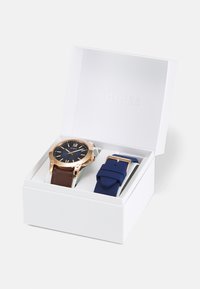 Goldfarbene Uhr mit dunkelblauem Ziffernblatt und braunem Lederarmband in einer weißen Box, neben einem zusätzlichen blauen Silikonarmband mit goldenen Beschlägen.