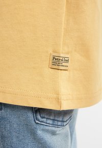 Gul bomulds T-shirt med et synligt label ved kanten. Syning detaljer langs den nederste kant. Båret med lyseblå denim jeans.