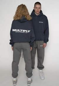 Felpa con cappuccio blu navy con logo "MULTIPLY" sulla schiena, abbinata a pantaloni della tuta grigi con logo sulla coscia, entrambi realizzati in tessuto morbido.