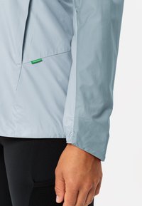 Lichtblauwe windbreaker met elastische manchetten, voorzien van een zijzak en een klein groen logotag. Gladde, lichte stoftextuur.