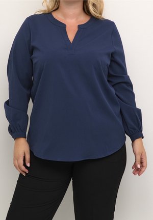 Kvinde iført en mørkeblå bluse med lange ærmer, delt udskæring og elastiske manchetter, kombineret med sorte bukser mod en ensfarvet baggrund.