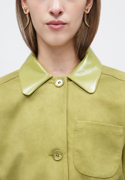 Γυναίκα που φοράει ανοιχτό πράσινο σακάκι από σουέντ με γυαλιστερό γιακά, κουμπιά και τσέπη στο στήθος, φορώντας επίσης χρυσούς κυρτούς σκουλαρίκια.