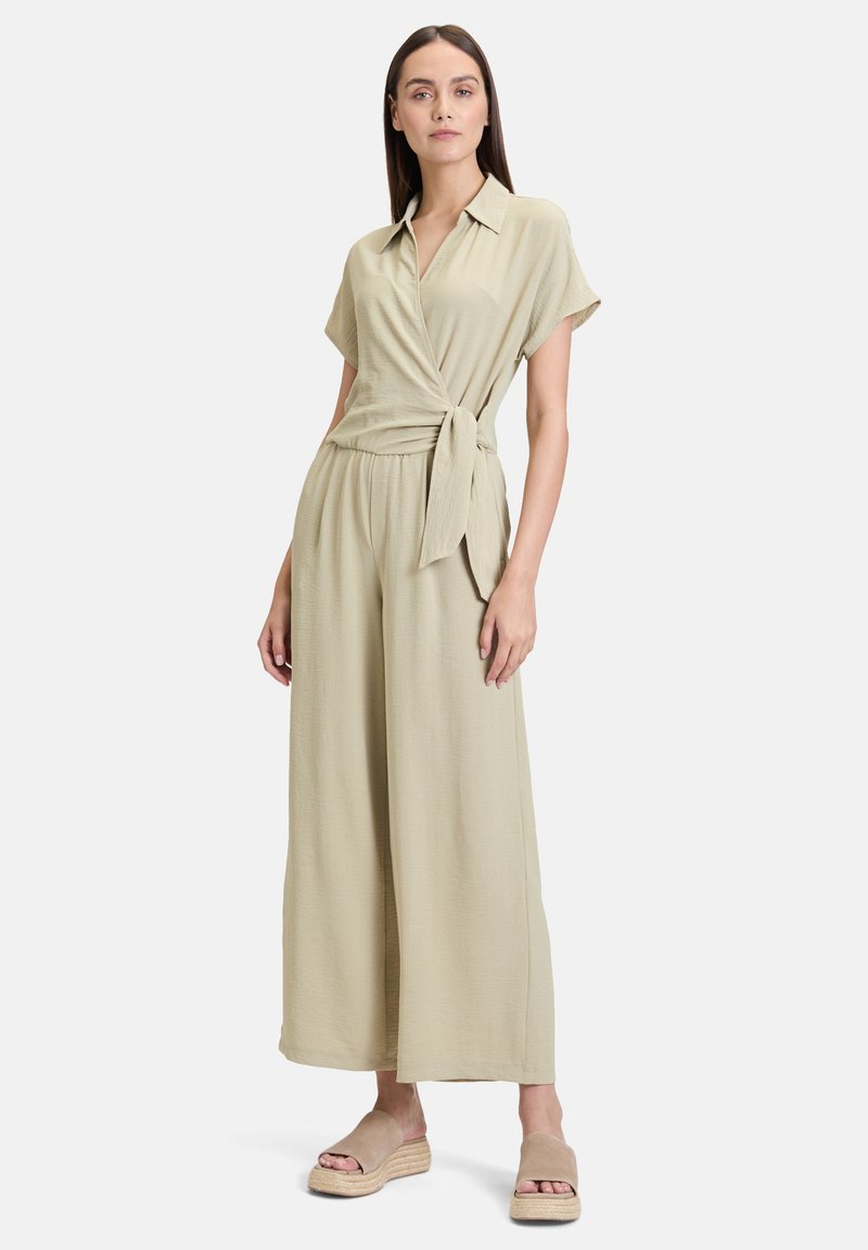 Lys beige wrap jumpsuit med korte ærmer, snøring i taljen og bred ben-designet, parret med beige platform sandaler. Glat tekstur.