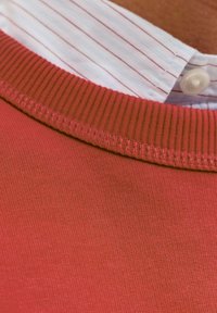 Czerwona bawełniana bluza z ribbowanym dekoltem, noszona na białej i różowej koszuli w paski z kołnierzykiem i zapięciem na guziki.