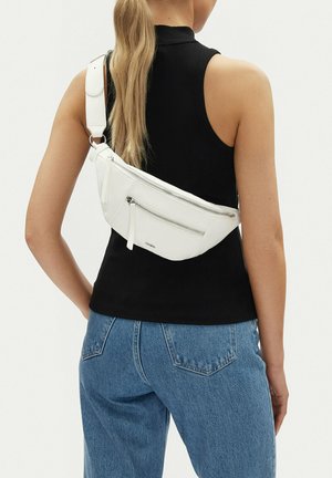 Žena s bílou koženou kabelkou přes rameno, oblečená v černém tílku a modrých džínách, zobrazená ze zadní strany.