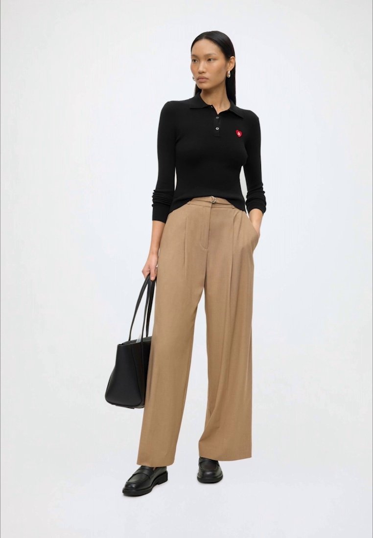 Femme aux longs cheveux foncés portant un polo noir à manches longues, un pantalon beige à jambes larges, des mocassins noirs, tenant un sac cabas noir, debout devant un fond uni.