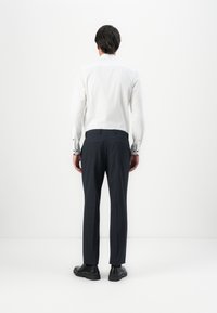 Man standing facing white wall, wearing white dress shirt, dark trousers, and black shoes.