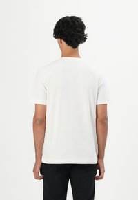 Mann mit lockigem, dunklem Haar, der ein einfaches, weißes, kurzärmliges Hemd und schwarze Hosen trägt, mit dem Rücken zu einem schlichten weißen Hintergrund.