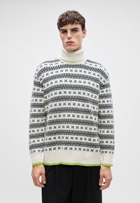 Halsgammel genser i hvit med svarte mønstre. Har ribbestrikkede mansjetter og nedkant, med en grønn detalj nederst.