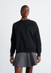 Sudadera negra con cuello redondo, puños acanalados y un pequeño parche de logo en el pecho, combinada con una falda gris con pliegues.