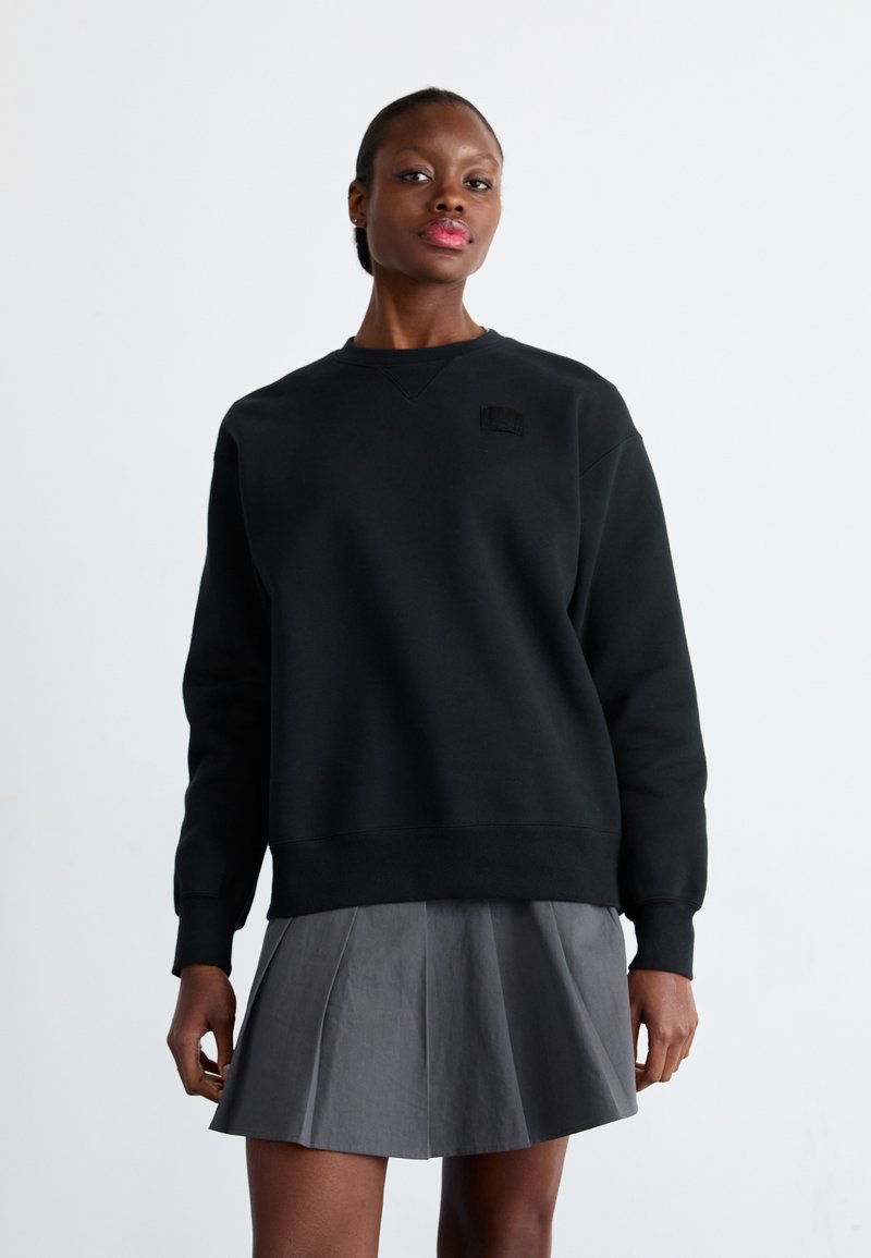 Sudadera negra con cuello redondo, puños acanalados y un pequeño parche de logo en el pecho, combinada con una falda gris con pliegues.