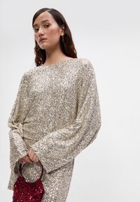 Šsparkly zlatasti top s sequinami in širokimi rokavi, ki ima teksturirano površino, v kombinaciji z majhno burgundsko torbico s krožno ročico.