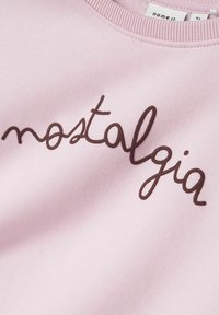 Tessuto rosa chiaro con testo cursivo marrone "nostalgia" e un'etichetta di abbigliamento visibile che legge "name it 4Y" vicino al colletto.