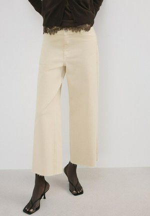 Mujer con pantalones anchos crema hasta la pantorrilla, sandalias negras de tacón con tiras, y una blusa oscura con encaje, de pie contra una pared blanca.