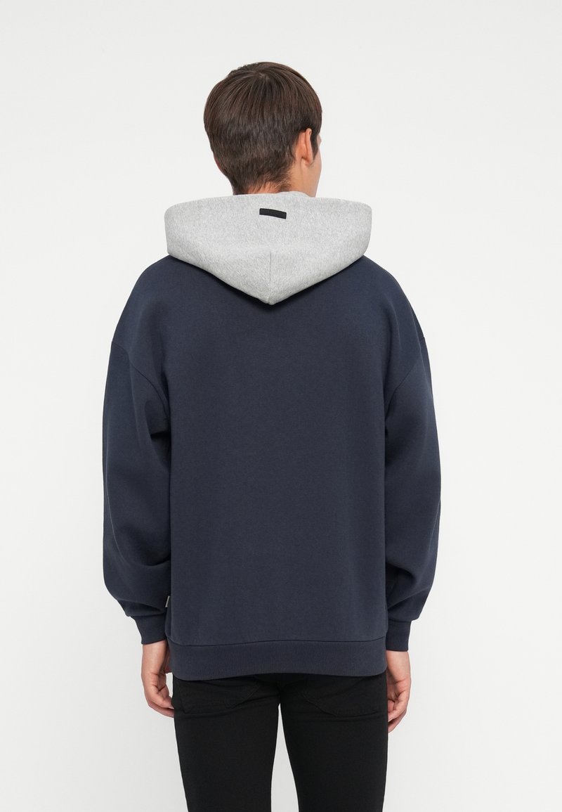 Marineblauer Hoodie mit einer hellgrauen gefütterten Kapuze. Er zeichnet sich durch eine lockere Passform, weichen Stoff und gerippte Bündchen aus. Auf der Rückseite ist ein minimalistisches Design zu sehen.