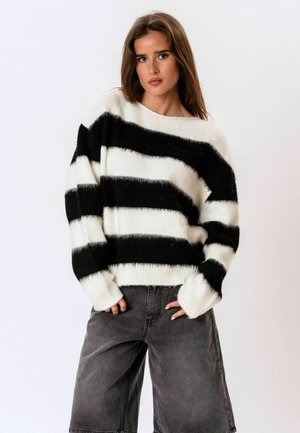 Jeune femme aux longs cheveux bruns portant un pull duveteux rayé noir et blanc et un short ample gris foncé sur un fond uni.