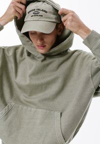 Hanorac de culoare verde măsliniu deschis, fabricat dintr-un material moale, cu o mare buzunar frontal, având o croială confortabilă și o glugă cu șnur.