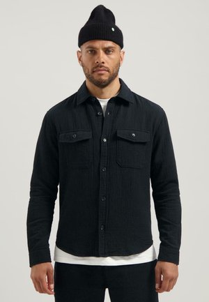 Man wearing a black textured button-up shirt over a white t-shirt with matching black pants and a black knit beanie against a plain background.