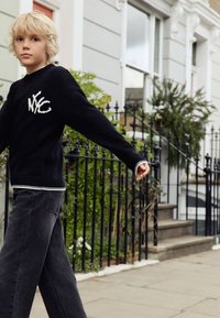 Černý pletený svetr s bílým nápisem "NYC", doplněný vybledlými černými širokými džínami. Má žebrovaný výstřih a rukávy. Neformální venkovní prostředí.