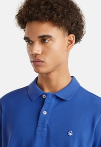 Camiseta polo azul con acabado texturizado, dos botones en el cuello y un pequeño logo bordado en la zona del pecho.