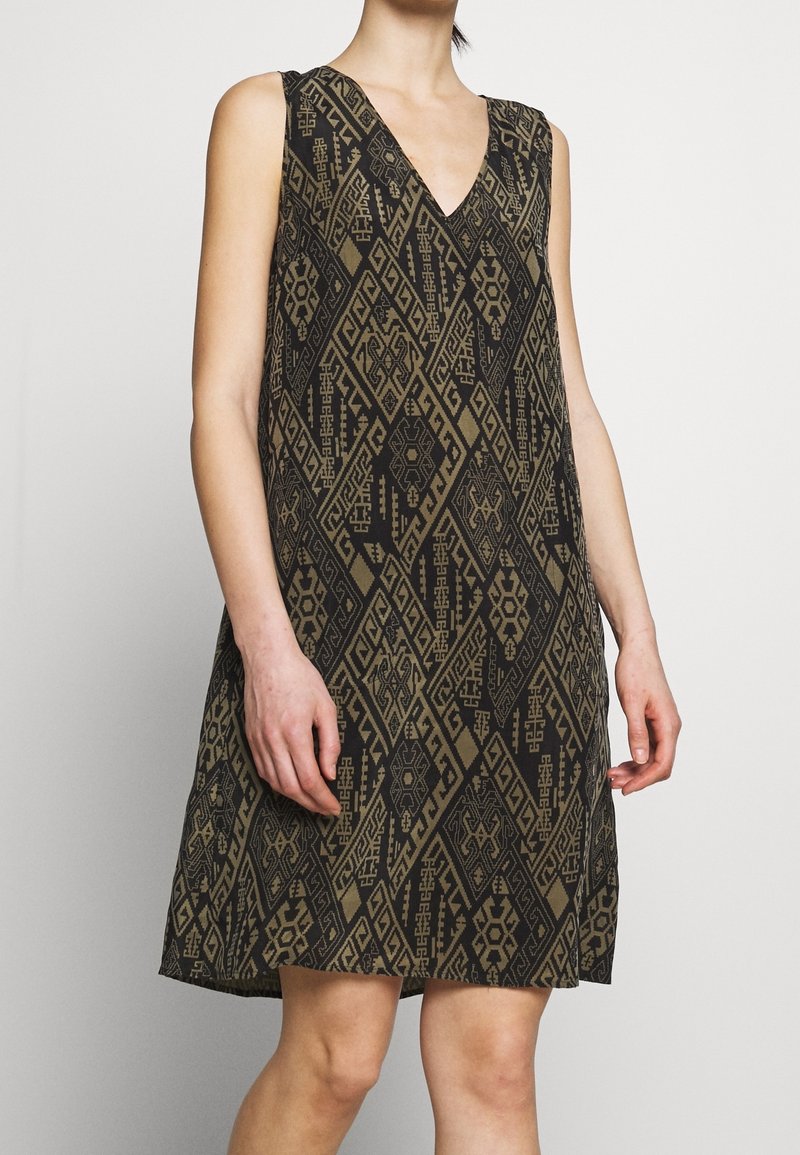 Ärmlös svart klänning med bruna geometriska tribalmönster, V-ringning, bärs av en person som står mot en enkel ljus bakgrund.