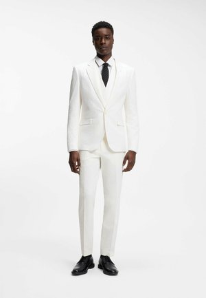 Man standing wearing a textured white suit with a white shirt, black striped tie, and black dress shoes against a plain white background.