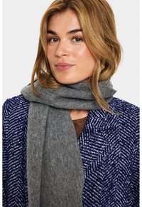 Kvinde med lysbrunt hår iført en blå og hvid mønstret frakke og et tykt gråt tørklæde, der kigger let til siden.