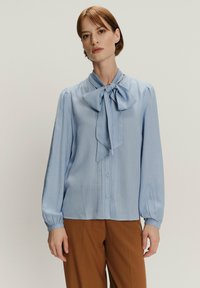 Jasnoniebieska bluzka z zawiązywanym kołnierzykiem w kształcie kokardy, pionowymi paskami, zapięciem na guziki i długimi, bufkami rękawami. Gładki materiał, casualowy krój.