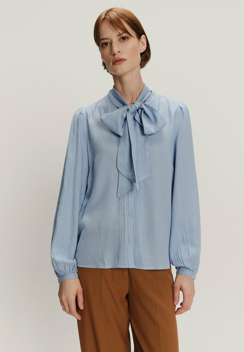Jasnoniebieska bluzka z zawiązywanym kołnierzykiem w kształcie kokardy, pionowymi paskami, zapięciem na guziki i długimi, bufkami rękawami. Gładki materiał, casualowy krój.