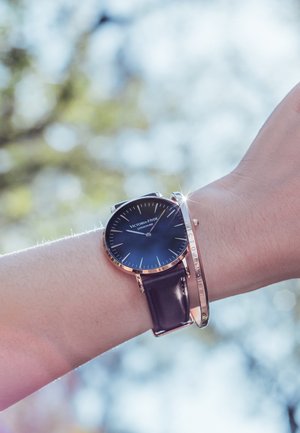 Montre avec bracelet en cuir noir et cadran rond bleu, marqueurs en ton argent et accents en rose doré poli ; associée à un bracelet métallique fin.