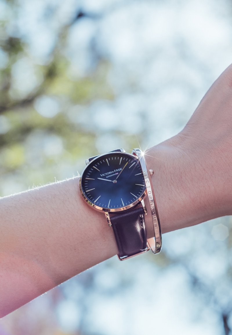 Reloj con correa de cuero negra y esfera redonda azul, marcadores en tono plateado y acentos en oro rosa pulido; acompañado de una pulsera metálica delgada.