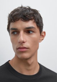 Giovane uomo con capelli corti e mossi scuri, che indossa una maglietta nera a collo rotondo, che guarda a sinistra su uno sfondo chiaro e semplice.