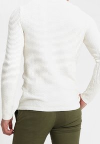 Weißer strukturierter Langarm-Strickpullover mit gerippten Details und taillierter Passform, kombiniert mit olivgrünen Hosen.