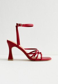 Rode sandalen met hoge hak en meerdere kruislings lopende banden, enkelband en een vierkante neus. Heeft een glanzende afwerking en een taps toelopende kegelhak.